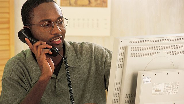 A man working on his computer while talking on the phone with a calendar on the wall behind him.