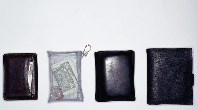 Four wallets lined up next to each other, three of them are dark leather and the fourth is clear plastic with money inside of it.