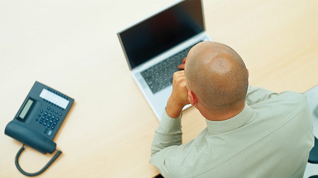 A man sitting by himself at a table with his laptop and an office phone.