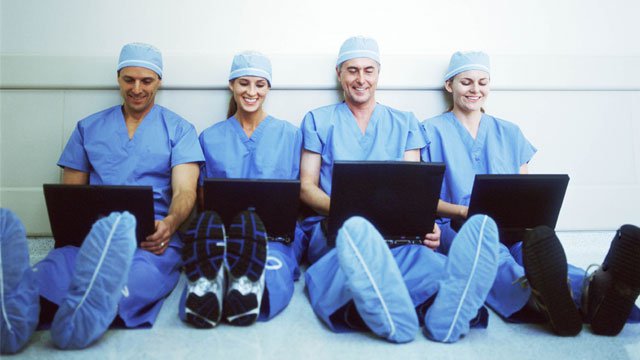 Four medical professionals wearing scrubs while sitting on the floor with their backs leaning up against the wall working on laptops.