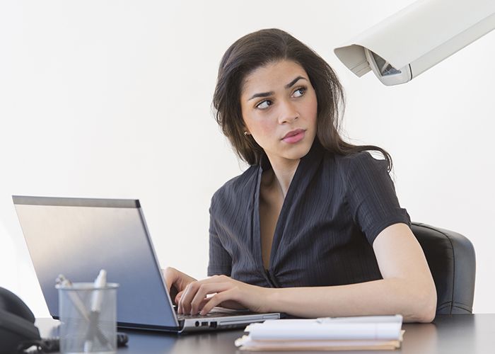 A woman on her computer looking over her shoulder under the watchful eye of a large security camera right next to her.