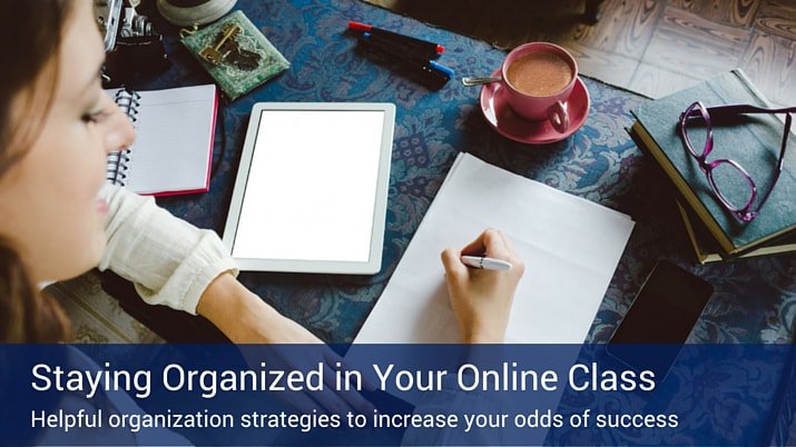 A woman writing on a piece of paper studying for her online classes with a tablet, coffee, notebook, glasses, and phone on a blue table.