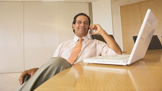 A man sitting at a table with a white laptop while talking on his cellphone.