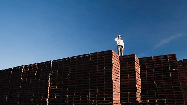 A man wearing a white coat and a hard hat standing on top of a tall stack of wooden pallets.