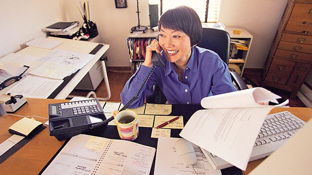A woman sitting at a cluttered desk full of paper while on her office phone looking at a sheet of paper.