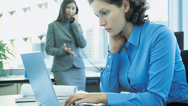 A woman at her desk typing on her computer with another woman in the background talking on an office phone.