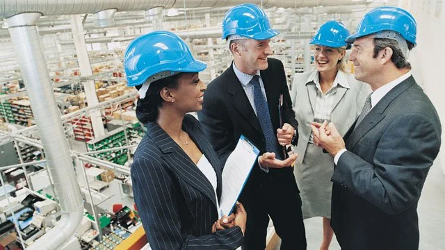 Four business professionals in a warehouse wearing blue hard construction hats.