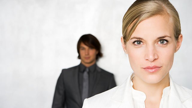 A close up headshot of a business woman wearing white, with a businessman wearing black stands against a white wall behind her.