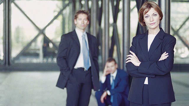 Three business people posing for a picture, with a woman standing in front with her arms crossed and two men standing behind her.