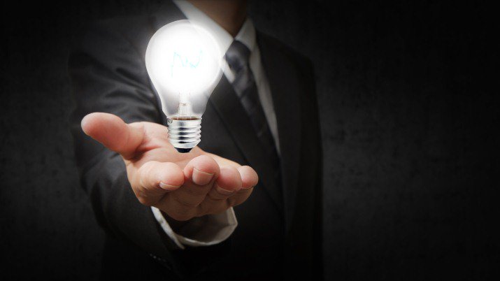 Close up of a man in a suit and tie holding out his hand with a light bulb floating in his hand.