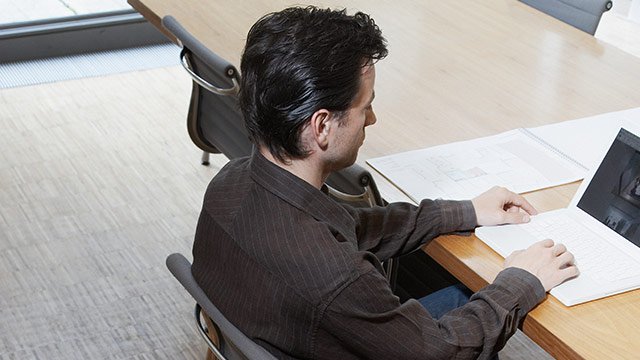 A student sitting by himself at a table working on his computer with papers laid out on the desk.