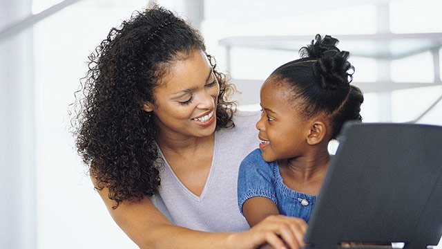A mother and her young daughter both looking at each other smiling while on the computer.