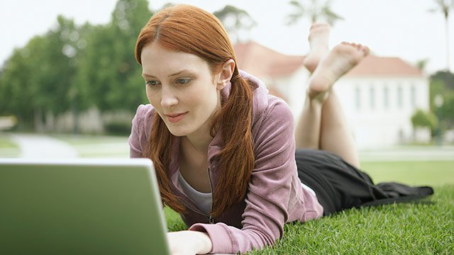 A woman laying on her stomach in the grass outside, working on IS security on her laptop.