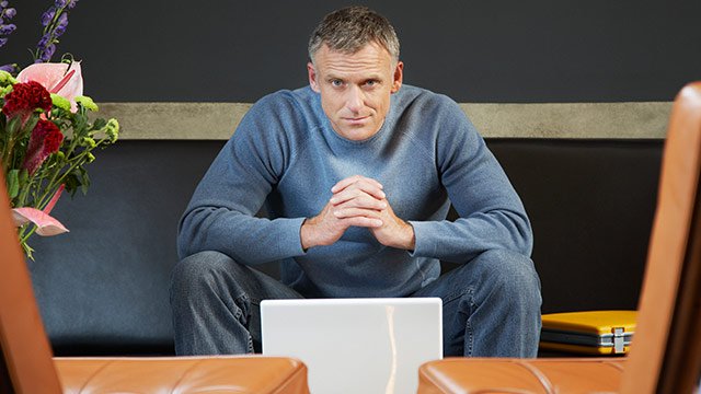 A man sitting down, working on his laptop looking into the camera.