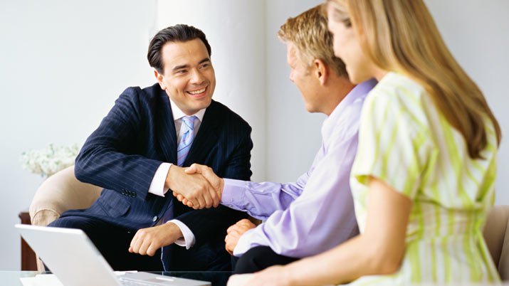 A meeting with a man wearing a business suit shaking hands with another man and a woman that are sitting together.