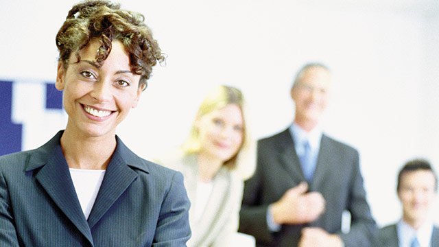 Project Manager depicted as a business woman smiling into the camera and three other co-workers behind her in the background.