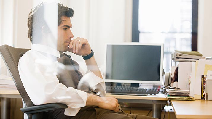 A man sitting in his office chair with a computer screen turned off behind him, thinking about the benefits of six sigma with his hand on his chin.