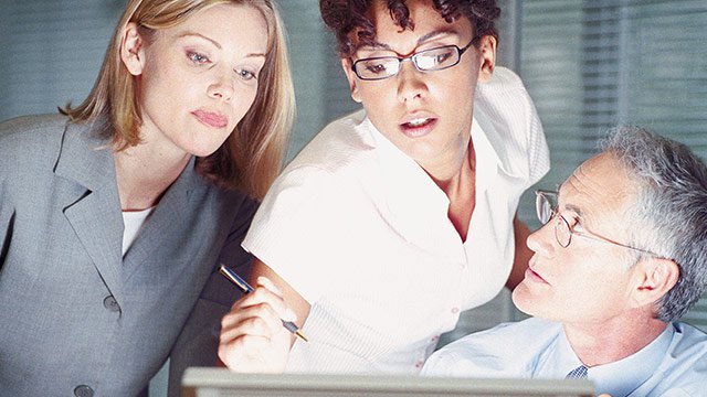 A man sitting at a computer with two women standing beside him helping each other figure something out on the computer screen.