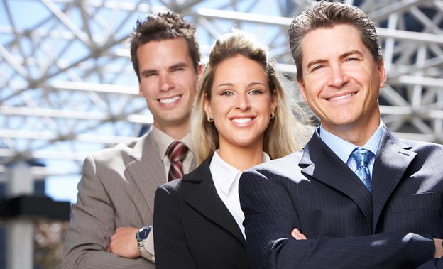 Three business people wearing suits, two men and one woman, smiling at the camera with a large artsy steel dome behind them.