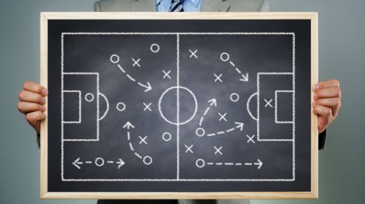 A person holding a small chalkboard with a soccer field drawing in chalk of x's and o's to represent a game plan.