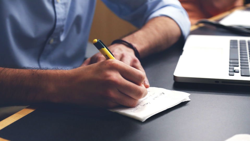 Close up of a man's hands writing on a piece of paper next to a laptop on a black desk.