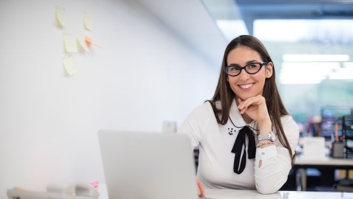 A scrummaster working on her computer with post it notes on the wall to help her plan the next agile sprint.