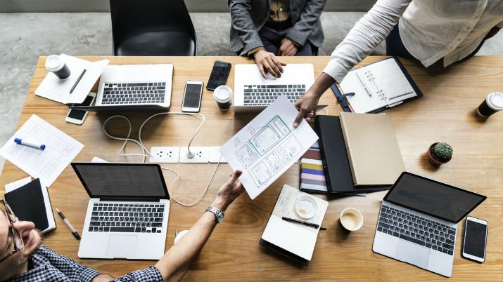 An overhead shot of a large cluttered desk with laptops, books, papers, phones, and coffee cups and two people handing paper to each other across the desk.