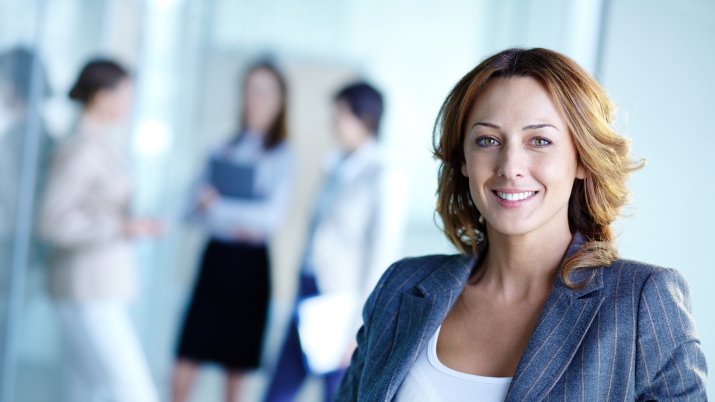 A business woman smiling into the camera with three other professionals out of focus in the background..