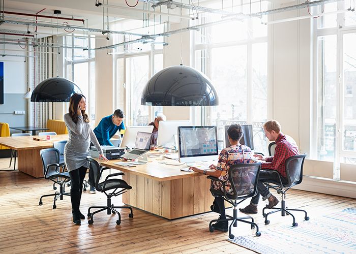 A modern office with large windows and five people sitting around a large desk with computers, working on managing virtual project teams.