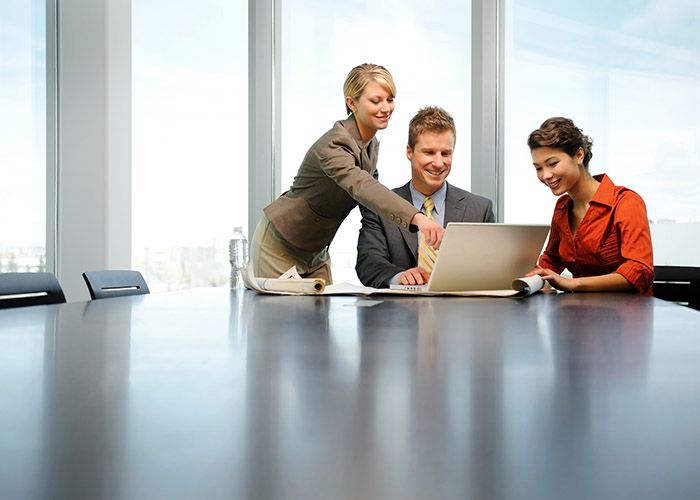 Three people in a highrise office at a board table all looking at a laptop with a window behind them overlooking a city.
