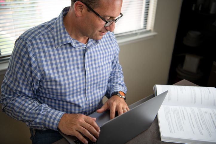 Adult learner at home reading textbook and using laptop.