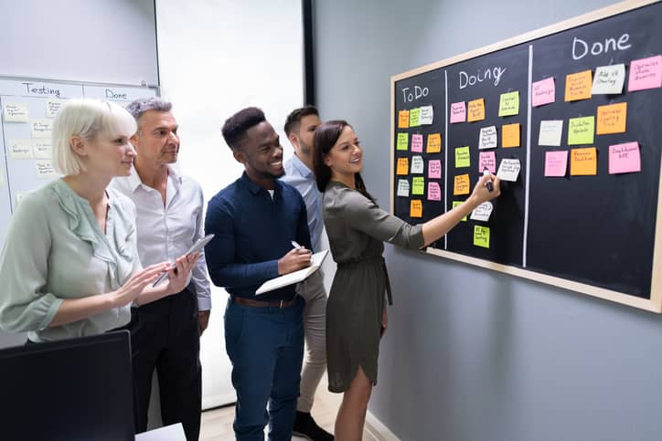 Diverse group of project team members standing in front of a sticky board looking at list of projects